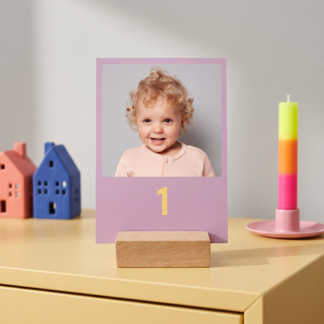 Erste Karte des Adventskalenders mit Fotos: Kind mit blonden Locken auf Lila, aufgestellt neben Keramikhäuschen und einer Kerze in Neonfarben